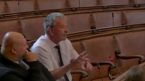 Wakefield Council Farmer has short grey hair and is seated at one of the desks and appears to be speaking. He is wearing a white shirt with a dark tie and is gesturing with one hand, with the fingers slightly extended as if emphasising a point. The active microphone with a red light is positioned just in front of this speaker, indicating that he is currently addressing the room.