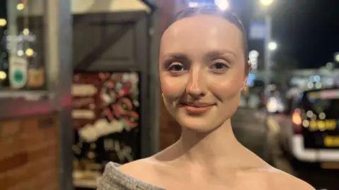 Elizabeth Baines / BBC A woman with her hair tied back smiles. Behind her a taxi on a rank is blurred.