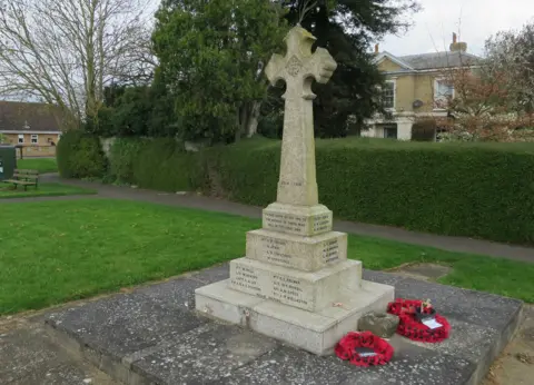 War memorial featuring a dedication and 13 names on three blocks of grey stone, with the largest stone at the base. A grey cross rises from the smallest stone at the top. All writing is too small to read. A wreath of red poppies has been laid to the right of the memorial, which is surrounded by a lawn and pathing, with hedges and trees beyond and a house and bungalow in the background. 