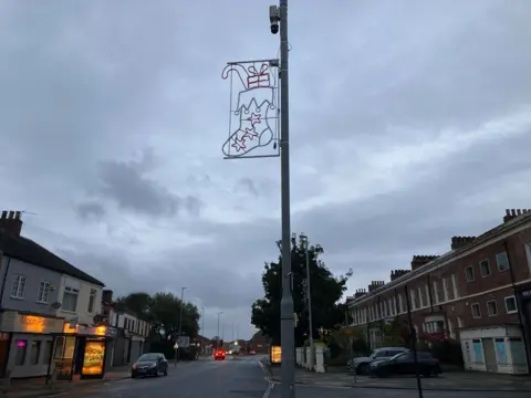 Gareth Lightfoot Christmas lights in the shape of a full Christmas stocking are attached to a CCTV post. A high street with various residences and shops can be seen in the background.