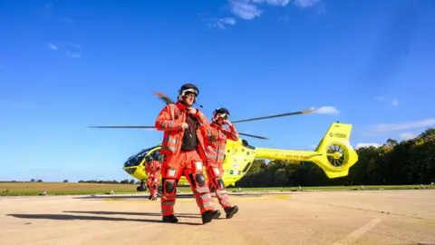 Yorkshire Air Ambulance Two people wearing high‑visibility orange protective flight suits and helmets walking across a concrete landing area. Behind them is a yellow helicopter with its rotor blades visible and the registration marking “G‑YORK” clearly readable on the tail section. The setting appears to be an open airfield or helipad, with flat paved ground, grassy fields in the distance, and a clear blue sky overhead. 