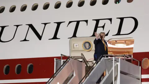 President Donald Trump is standing at the top of a set of steps leading to the door of a large plane. He is wearing a blue suit and is waving.