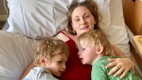 Charlotte Coulbert A woman in a bed hugging two children who are lying on top of her.