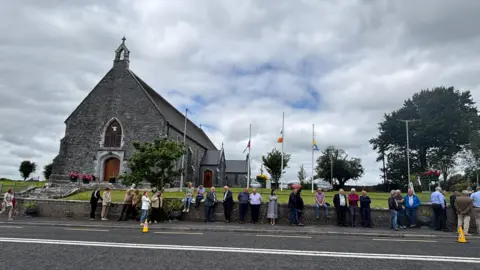 People lined up on a street outside a church. There is a cloudy sky above.