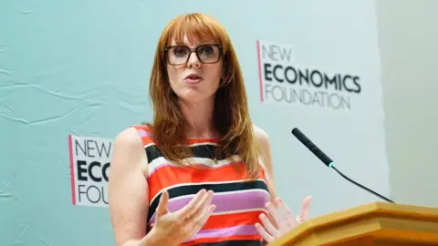 PA Media Angela Rayner in a black, white, orange and lilac striped dress and wearing brown-framed glasses is standing at a lectern 