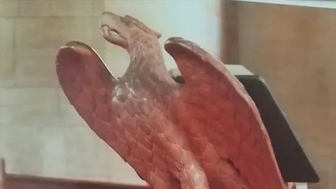 A wooden eagle animal carving atop a lectern, with an open book on its back 