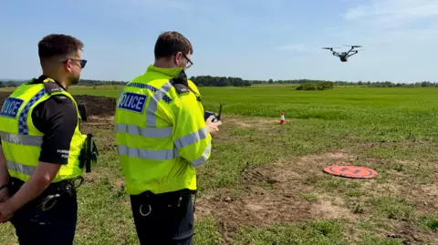 Two male police officers in hi-vis uniforms operate an aerial drone from a small landing pad in a field. It is a bright sunny day and one of the officers wears sunglasses.
