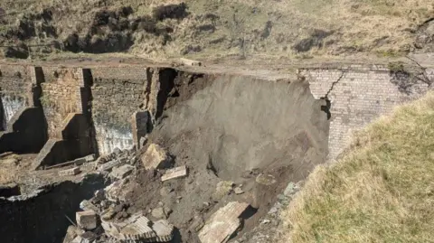 A large wall has collapsed. There is rubble and a large mudbank that has flowed out of it. The remainder of a wall is still standing on either side.
