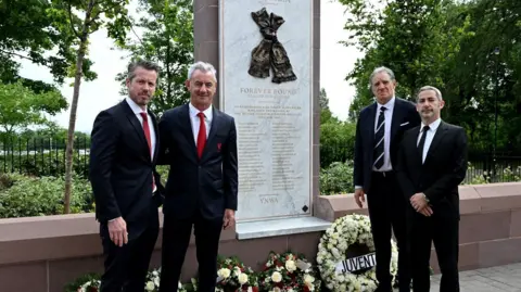 LFC Four men dressed in black suits stand in front of the memorial where wreaths have been laid
