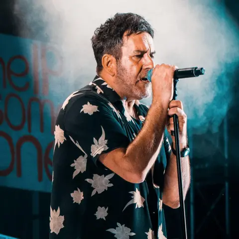 Sterling Chandler Terry Hall is wearing a dark shirt with white flowers on it. He is singing into a microphone during the 2019 concert at Coventry Cathedral 