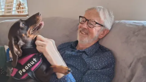 Supplied Tim Strugnell sitting on a sofa while wearing a shirt and glasses. There is a small brown dog on his lap which he is stroking. He is smiling and seems happy.
