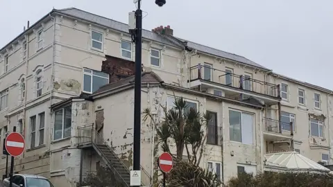 Isle of Wight Council Rear view of large sprawling cream painted hotel, many of its walls have peeling paint and missing render - a road runs to the left and plants are growing along the rear.