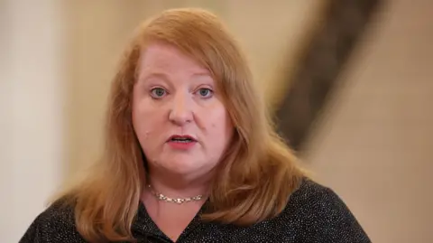 PA Media Naomi  Long. She has shoulder-length strawberry blonde hair, wearing a black blouse and silver necklace.