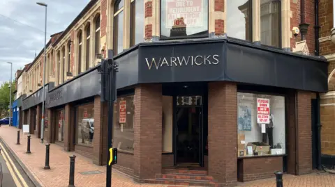 The outside of Warwicks now. It has closing down sales in the window, clothes in the main windows, and the front is made of brick, with a metal frontage for the sign in black. Bollards are outside the shop. There are steps up to the building. 
