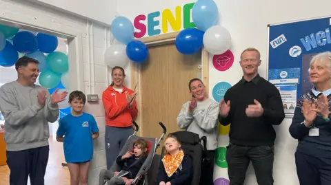 BBC Left to right, a man in a grey jumper and blue trousers, a boy in an oversized blue top and short black shorts, a woman in an orange hoodie and grey trousers, a woman in a grey jumper and black trousers, a man in a black top, jumper and trousers and a woman in navy blue jumper and trousers. They are all clapping. In front are two children in pushchairs. Behind them on the wall are blue balloons. There is a brown door and a sign saying "SEND" above it.