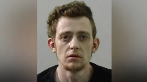 Northumbria Police Mugshot of Reece Galbraith. He has thick ginger hair and a ginger goatee and a very pale face. He is wearing a black t shirt