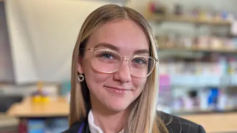 Erin, with blonde hair and glasses, sits in her classroom