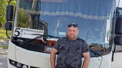 A man stands in front of a bus with the number 5 on the front wearing sunglasses and a grey t-shirt.