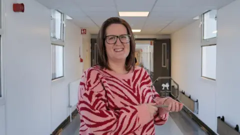 West Suffolk Foundation Trust Woman wearing a pink and red animal print jumper holds an award-a sculpture made of glass and she stand in a hospital corridor. 