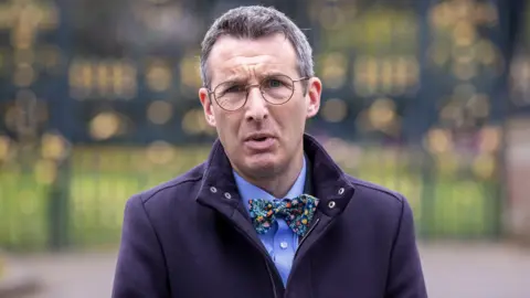 Andrew Muir is standing outside on front of large black gates. He wears glasses and is wearing a navy coat with a blue shirt and a bow tie.