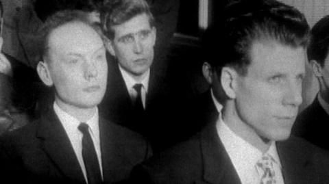 Black and white image of three young well-dressed men in jackets and ties, with their gaze fixed over the right shoulder of the camera towards where they are being spoken to from.