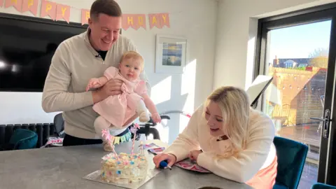 Laura's husband hold Aurora over her birthday cake over a kitchen counter has her mum lights the council. The family are smiling with birthday decorations seen in the background.