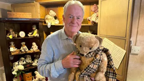 A man holding a beat that is wearing a scarf. He is standing in front of a cabinet with more stuffed toys on the shelves