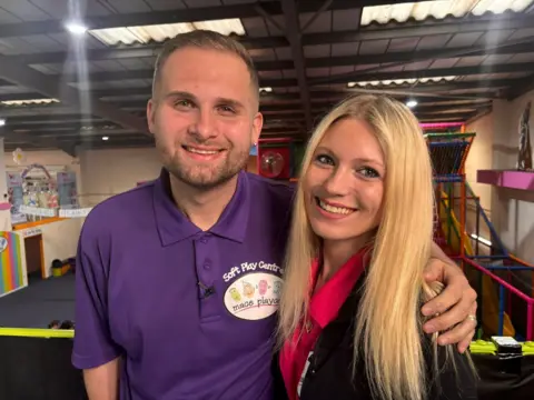 Boston wearing a purple soft play centre branded polo shirt and smiling with his arm around his sister's shoulders. She has long straight blonde hair and is also smiling wearing a red polo shirt with the same branding and a black cardigan
