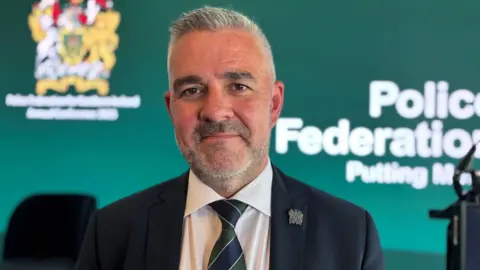 Liam Kelly, a man with grey hair and stubble, wearing a black jacket, white shirt and blue and green striped tie. There is a green backdrop with a white Police Federation logo on it.