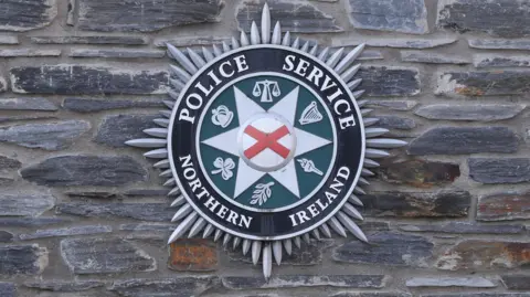 A PSNI badge is on a brick wall. The badge has the words 'Police Service of Northern Ireland' in white writing with a red cross in the middle and harps, scales, crowns and leaves on green background.