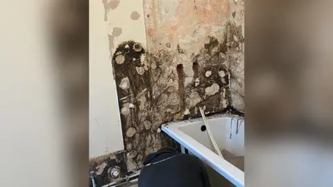 Tiles have been removed in a bathroom revealing dark mould on the wall behind a white bathtub