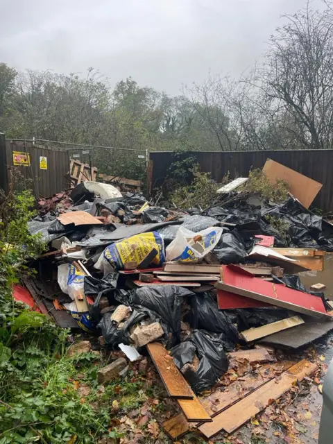 Buckinghamshire Council A large pile of waste dumped in front of a fence, including filled black bin bags and planks of wood.