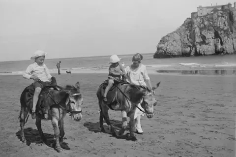Getty Images A woman leads walks alongside two donkeys carrying small children on Tenby's South Beach in 1924 in a black and white photo. Shewears a long white dress. The children are wearing shorts, shirt and sunhat and a dress and a sunhat. A man and a dog are visible in the background at the water's edge.