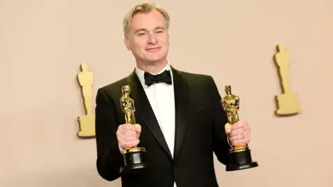 Getty Images A smiling man wearing a tuxedo and holding his two Oscars in his hands.