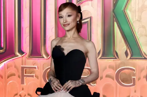 Reuters Ariana Grande wears a black and white dress and stands in front of a billboard promoting the film Wicked: For Good. 