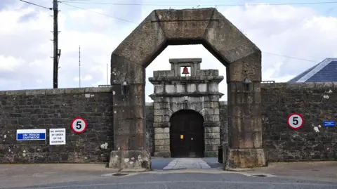 The entrance to HMP Dartmoor in Devon. It consists of a large stone arch and a large stone wall. Another archway with a door in it can be seen through the other arch.