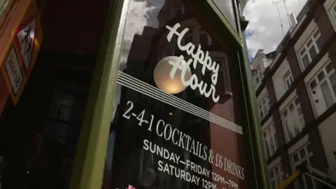 Sign on a pub winder reading Happy Hour in white writing on a brown background with the camera pointing up to the sky