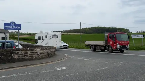 A crossroads with traffic travelling along the main road. A blue sign for "Paddle School" is visible on the left, inside the school grounds' stone boundary wall, which runs along the corner of the junction.