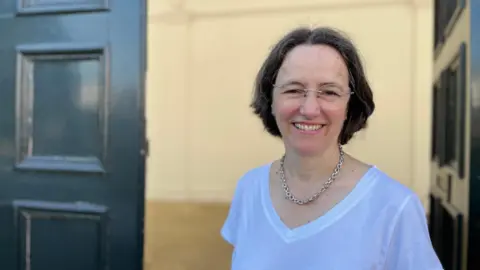 Tom Jackson/BBC Head and shoulders of Karen Young. Karen has dark shoulder length hair, is wearing glasses and a chain-like necklace and white top. She is standing in an open doorway.