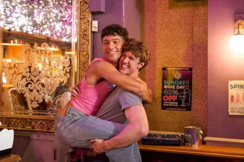 Channel 4 / James Stack Jackson Connor has his legs wrapped around Joseph Evans' waist with both men laughing in a bar. Jackson wears a pink vest and jeans and Joseph wears a grey t-shirt 