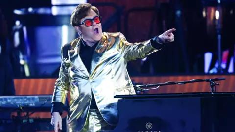 Elton John points out at the crowd on stage. He is wearing large glasses with bright red lenses and a gold suit.