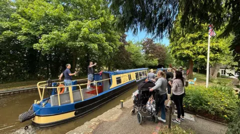 Derwen College A blue and yellow boat on a canal with a number of people, one of them in a wheelchair, waving from the towpath