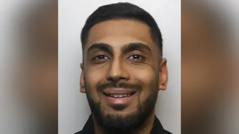 A police mugshot of Uzair Hamid. He has short dark hair and dark facial hair.