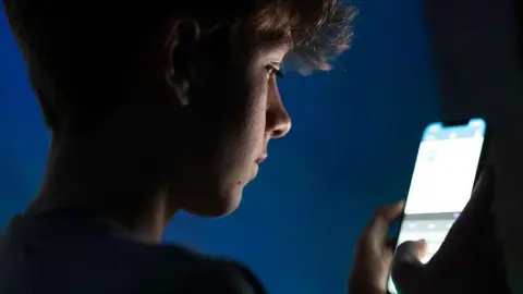 A teenage boy looks at an iPhone screen displaying various social media apps. It is dark in the room.