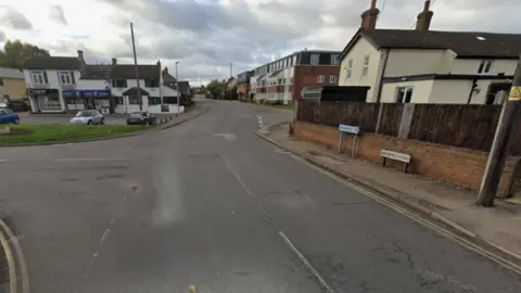 Greenfield Road leading into Flitwick, with houses and small shops on either side of the road and double yellow lines.