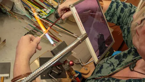 Pete Robinson A close-up picture of a woman working with a blow torch to create glass beads