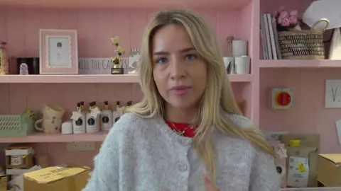 Promise Fulstow is a shop assistant at gift shop, U Melt Me, and is pictured behind the counter next to gifts and packaging and has blonde hair and wears a wool cardigan. 