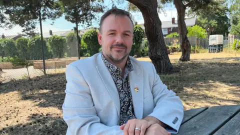 Clare Worden/BBC A man looks at the camera. He is wearing a light blue jacket and grey and black patterned shirt. He has short brown hair and a beard. 