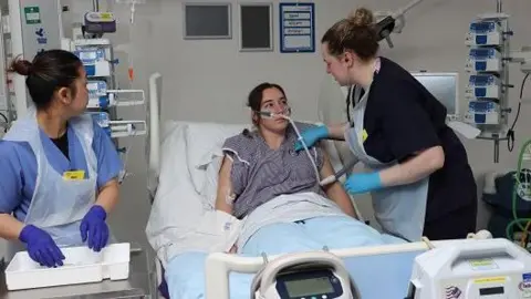 A young woman with a tube attached to her face being treated by two nurses while lying on a hospital bed surrounded by medical equipment. 