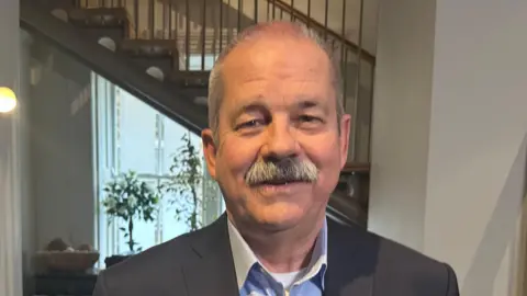 Rob Drury is wearing a suit and a shirt with no tie. He has grey hair and a grey moustache. He is in a hotel lobby and is smiling.